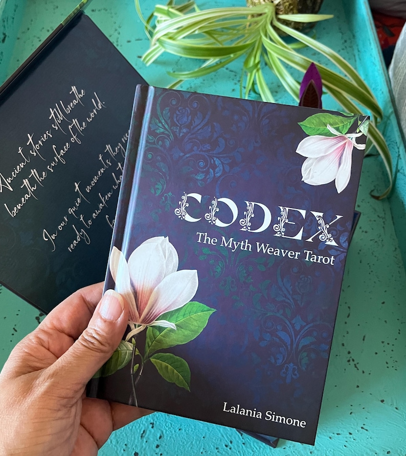 May include: A hardback book titled "CODEX The Myth Weaver Tarot" with a dark blue cover featuring a floral design and a white magnolia. The author's name, Lalania Simone, is at the bottom. Another book with text is in the background.