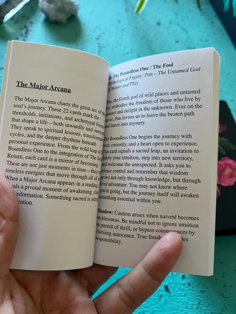 May include: An open book with text about "The Major Arcana" and "The Boundless One / The Fool" tarot card. The text explains the card's meaning, focusing on spiritual lessons and personal growth. The book is held open, revealing the pages.