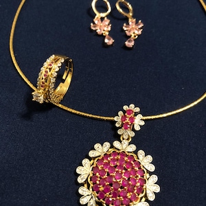 May include: A gold-toned jewelry set featuring a ring, earrings, and a necklace. The ring has a central clear stone and a band with red and clear stones. The earrings and necklace pendant have pink and clear stones in a floral design.