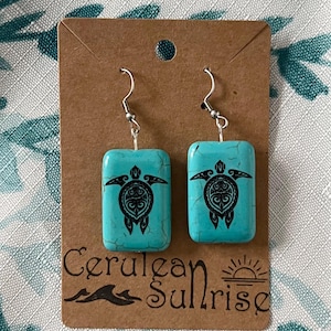 May include: Turquoise rectangular earrings with a black turtle design. The earrings are displayed on a brown card with the text "Cerulean Sunrise" and a sun graphic. The earrings have silver hooks.
