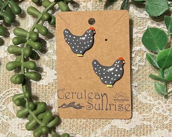 May include: A pair of ceramic chicken earrings with black and white polka dots and orange combs. The earrings are displayed on a brown card with the text "Cerulean Sunrise" and a stylized sun and waves.