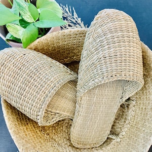 May include: Two woven straw slippers in a woven straw basket. The slippers are light brown and have a simple design.