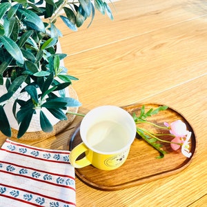 May include: A yellow ceramic mug with a bee design sits on a wooden tray with pink flowers. A white and red striped tea towel is in the foreground.