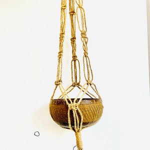 May include: A brown coconut shell planter hanging from a natural fiber macrame plant hanger. The hanger has a tassel at the bottom. The planter is hanging against a white wall with a child's drawing of a snowman and a reindeer on it.