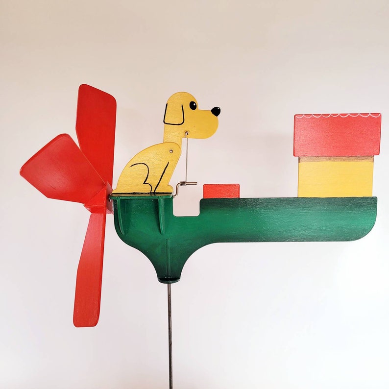 May include: A wooden wind spinner featuring a yellow dog sitting on a green base. The dog is looking to the right. The wind spinner has a red propeller with three blades. The base has a small red box and a yellow house on top.