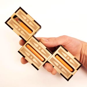 Puede incluir: Tres cajas rectangulares de madera con bordes oscuros y superficies de madera clara, cada una con una ranura central que contiene un lápiz naranja. Las cajas están dispuestas en forma de escalera y muestran recortes geométricos.