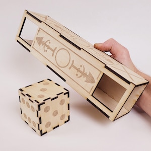 Puede incluir: Una caja de dados de madera y una caja rectangular con diseños grabados. La caja de dados es un cubo con marcas de puntos circulares. La caja rectangular tiene un diseño grabado de un círculo y flechas. Ambos artículos están hechos de madera clara.