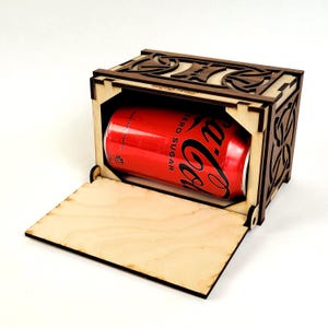 Op de afbeelding: Een lasergesneden houten kist met een rode Coca-Cola Zero Sugar-blikje. De kist heeft decoratieve uitsparingen en een open deur. De kist is gemaakt van licht hout met donkere accenten. Het blikje zit in de kist.