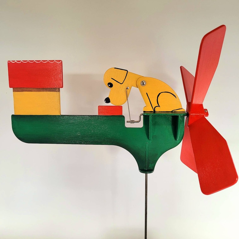 May include: A wooden wind spinner featuring a red, yellow and green colour scheme. A yellow dog figure is attached to the spinner and is looking down at a small red box. The spinner has three red blades.