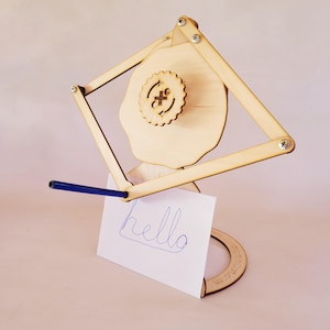 May include: A light-colored wooden mechanical drawing machine featuring a diamond-shaped linkage frame, a central gear with a plus sign and circular arrows, and a blue pen attached to the arm, holding a white card with the handwritten word hello.