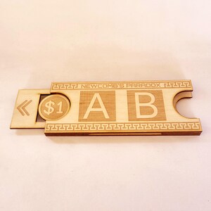 Puede incluir: Juego de mesa de madera con el texto "NEWCOMB'S PARADOX" y las letras "A" y "B". Una ranura contiene una moneda de madera con el símbolo "$1". El tablero tiene un borde decorativo y un símbolo de flecha.