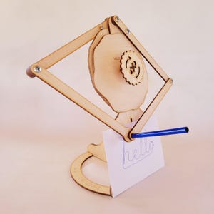 May include: A light-colored wooden pantograph drawing machine featuring a central gear mechanism with a plus sign symbol. The device includes a blue pen attached to the arm, which is currently holding a white card with the word hello written on it.