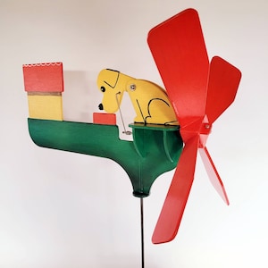 May include: A wooden wind spinner featuring a yellow dog figurine and red and green painted details. The dog is perched on a green platform and the wind spinner has four red blades.