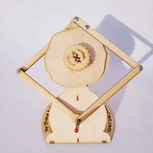 May include: A light-colored wooden mechanical drawing device featuring a central gear with an X symbol and circular arrows, connected to a diamond-shaped frame with metal screws. The base includes the text by Shasa and the website address.