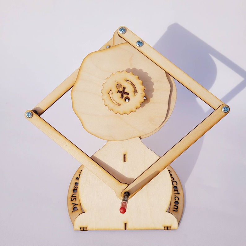 May include: A light-colored wooden mechanical drawing device featuring a central gear with an X symbol and circular arrows, connected to a diamond-shaped frame with metal screws. The base includes the text by Shasa and the website address.