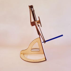 May include: A light-colored wooden stand with a curved base and a triangular cutout, supporting a vertical frame assembly with metal fasteners. A thin blue pen is inserted into the side of the frame, extending outward from the structure.