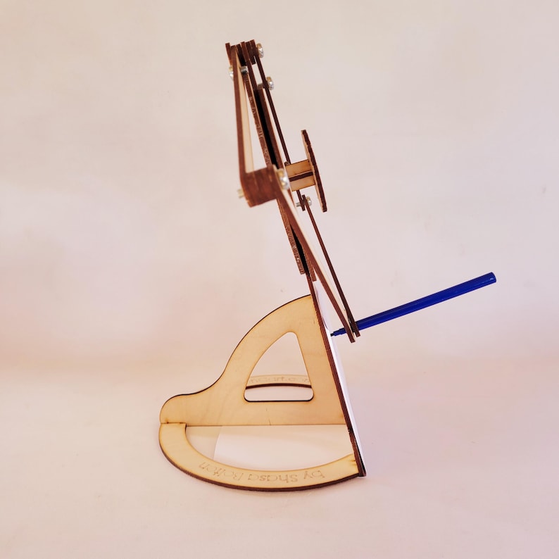 May include: A light-colored wooden stand with a curved base and a triangular cutout, supporting a vertical frame assembly with metal fasteners. A thin blue pen is inserted into the side of the frame, extending outward from the structure.