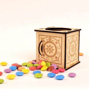 Puede incluir: Una caja cuadrada de madera con una abertura circular en la parte superior y un asa lateral, con patrones florales cortados con láser en sus paneles. La caja está rodeada por una dispersión de pequeños dulces de chocolate redondos y coloridos.
