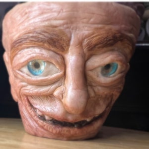 May include: A ceramic planter sculpted in the form of a human face, featuring a broad, grinning mouth. The face is painted in brown and tan hues, with blue eyes. The planter has a textured finish, suggesting a handmade quality and a unique design.