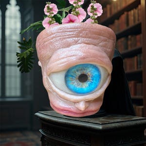 Può includere: Un vaso da fiori unico a forma di occhio umano con un'iride blu e un esterno carnoso rosa. Il vaso è pieno di fiori rosa.