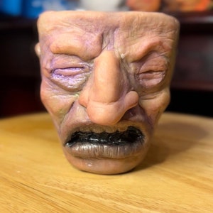 May include: A ceramic planter shaped like a face with a furrowed brow, closed eyes, and a wide open mouth. The planter is painted in shades of brown, beige, and purple.