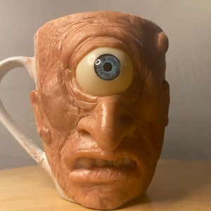 May include: A ceramic coffee mug shaped like a one-eyed monster with a blue eye. The mug has a white handle and a textured, skin-like surface.