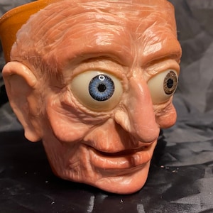 May include: A distinctive planter sculpted in the form of a human head. The piece showcases realistic skin tones, detailed wrinkles, and expressive eyes with blue and brown irises. The top of the head is open for planting.