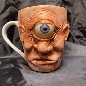 May include: A ceramic coffee mug shaped like a face with a single large blue eye. The mug has a white handle and a textured surface.