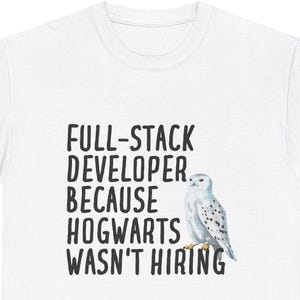 Op de afbeelding: Wit T-shirt met de tekst "FULL-STACK DEVELOPER BECAUSE HOGWARTS WASN'T HIRING" in zwarte letters. Een aquarel illustratie van een witte uil met zwarte vlekken staat rechts van de tekst. Dit is een casual kledingstuk.