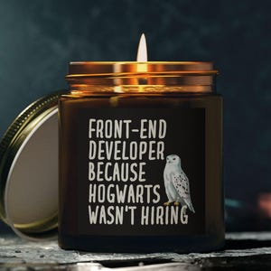 Könnte beinhalten: Eine brennende Kerze in einem braunen Glas mit einem goldenen Deckel. Das Glas hat ein Etikett mit dem Text "FRONT-END DEVELOPER BECAUSE HOGWARTS WASN'T HIRING" und einer Zeichnung einer weißen Eule. Der Deckel ist teilweise geöffnet.