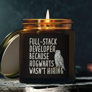 Peut inclure: Une bougie allumée dans un pot en verre marron avec un couvercle doré. L'étiquette indique "FULL-STACK DEVELOPER BECAUSE HOGWARTS WASN'T HIRING" avec une illustration de hibou blanc. Le pot est ouvert, et le fond est sombre.