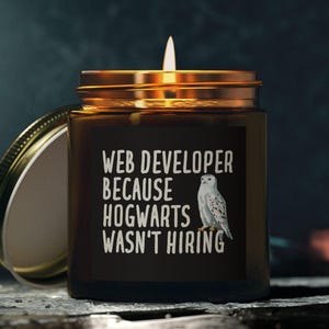 May include: A lit candle in a brown glass jar with a gold lid. The label reads "WEB DEVELOPER BECAUSE HOGWARTS WASN'T HIRING" with a white owl illustration. The lid is off and resting beside the jar.