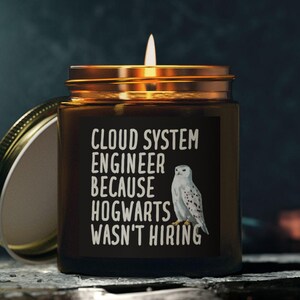 May include: A lit candle in a brown glass jar with a gold lid. The label reads "CLOUD SYSTEM ENGINEER BECAUSE HOGWARTS WASN'T HIRING" with a white owl illustration. The lid is off to the side.