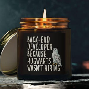 May include: A lit candle in a brown glass jar with a gold lid. The jar has the text "BACK-END DEVELOPER BECAUSE HOGWARTS WASN'T HIRING" and a white owl illustration. The lid is off, and the candle is on a dark surface.