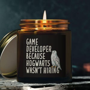 May include: A lit, dark brown candle in a glass jar with a gold lid. The candle has white text that reads "GAME DEVELOPER BECAUSE HOGWARTS WASN'T HIRING" with a white owl graphic. The lid is off and resting next to the jar.
