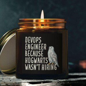 May include: A lit candle in a brown glass jar with a gold lid. The jar features the text "DevOps Engineer Because Hogwarts Wasn't Hiring" and a white owl illustration. The lid is off and resting beside the jar.
