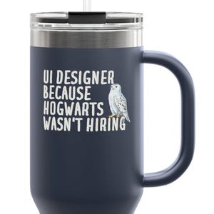 May include: Navy blue travel mug with a handle and a white straw. The mug features the text "UI DESIGNER BECAUSE HOGWARTS WASN'T HIRING" and a graphic of a white owl.