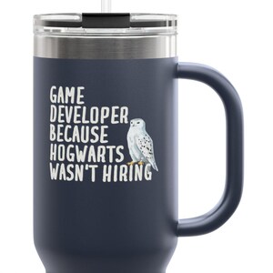 May include: Navy blue travel mug with a handle and a white straw. The mug features the text "GAME DEVELOPER BECAUSE HOGWARTS WASN'T HIRING" and a white owl illustration. The mug is designed to keep beverages hot or cold.