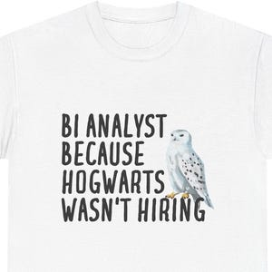 May include: White t-shirt with the text "BI ANALYST BECAUSE HOGWARTS WASN'T HIRING" in black font. A watercolor illustration of a snowy owl is to the right of the text. The t-shirt is a casual wear item.