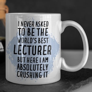 Könnte beinhalten: Eine weiße Keramik-Kaffeetasse mit einem blauen und weißen Spritzmuster. Die Tasse hat eine schwarze Aufschrift mit dem Text "Ich habe nie darum gebeten, der beste Dozent der Welt zu sein, aber hier bin ich und rocke es."