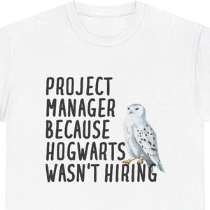 Puede incluir: Camiseta blanca con el texto "PROJECT MANAGER BECAUSE HOGWARTS WASN'T HIRING" en fuente negra. A la derecha del texto hay una ilustración de acuarela de un búho nival. La camiseta es una prenda informal.