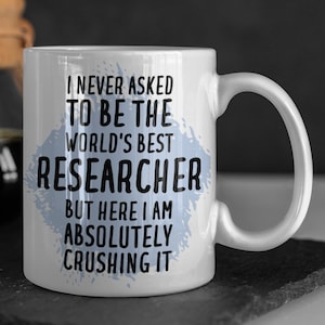 May include: White ceramic mug with a blue and white design. The mug has black text that reads "I never asked to be the world's best researcher but here I am absolutely crushing it."