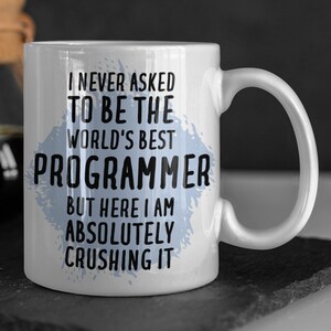 May include: White ceramic mug with a blue and white splatter design. The mug has black text that reads "I never asked to be the world's best programmer but here I am absolutely crushing it."