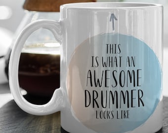 Op de afbeelding: Een witte keramische mok met een blauw en roze cirkelontwerp. De tekst op de mok luidt "This is what an awesome drummer looks like".