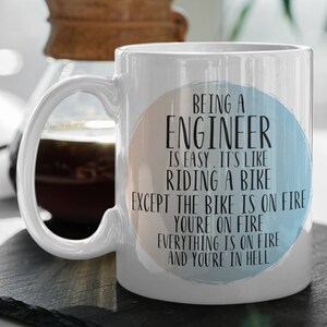 May include: White ceramic mug with a blue and orange watercolor circle design. The text on the mug reads "Being an Engineer is easy. It's like riding a bike except the bike is on fire, you're on fire, everything is on fire and you're in hell."