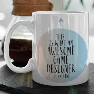 May include: White ceramic coffee mug with a blue and peach watercolor circle design. The text on the mug reads "This is what an awesome game designer looks like".