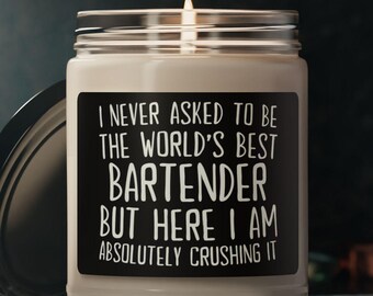 May include: A white candle in a glass jar with a black label. The label reads, "I NEVER ASKED TO BE THE WORLD'S BEST BARTENDER BUT HERE I AM ABSOLUTELY CRUSHING IT." The candle has a silver lid and a lit wick.