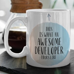 Op de afbeelding: Een witte keramische koffiemok met een blauw en perzikkleurig cirkelontwerp. De mok heeft een zwart handvat en de tekst "This is what an awesome developer looks like" gedrukt op de cirkel.