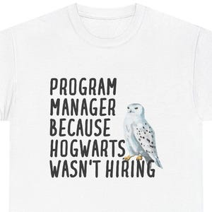 Puede incluir: Camiseta blanca con el texto "PROGRAM MANAGER BECAUSE HOGWARTS WASN'T HIRING" en negro, acompañada de una ilustración de acuarela de un búho nival. Prenda de vestir informal.