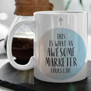 May include: White ceramic coffee mug with a blue and peach watercolor circle design. The text "This is what an awesome marketer looks like" is printed in black on the mug.